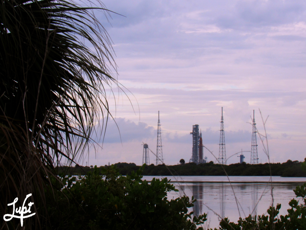 A cotton candy sunset sky frames the scene, lavender and pink as it reflects in the water. A fair amount of scrub and trees lay at the base of the image, a lone palm climbing the left side of the frame. Clear across the water, and smoothly reflected within, sits the SLS rocket and pad 39b, the marsh waters like frosted glass.