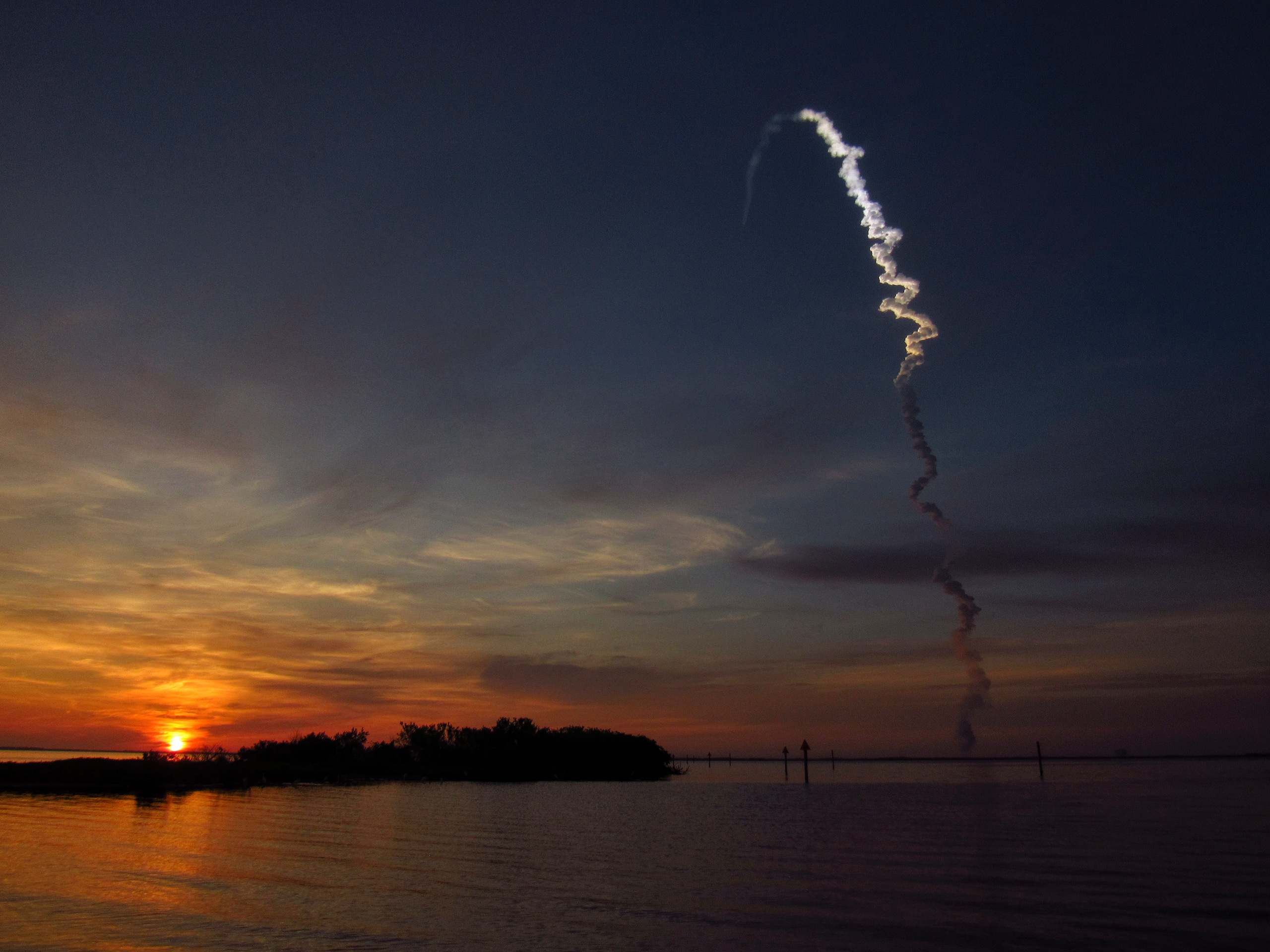 The sun just barely peeks above the horizon, giving the bottom left corner of the frame a rich orange glow as it fades into the gunmetal blues and indigos of a steely morning sky, daubbed with wispy clouds. The smoke trail of an Atlas V rocket ascends from the opposite end of the horizon, first cast in shadowed grey and then taking on the bright hues of daylight as it climbs into the morning sun, arcing into the distance.